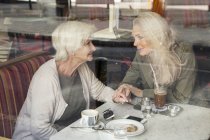 Madre e hija sentadas juntas en la cafetería, cogidas de la mano, vistas por la ventana de la cafetería - foto de stock