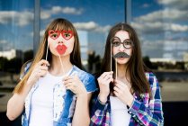 Portrait of two young women holding up lips and glasses costume masks — Stock Photo