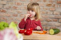 Cute toddler girl in kitchen eating raw vegetables — Stock Photo