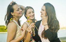 Three young female friends on riverbank holding dandelions — Stock Photo