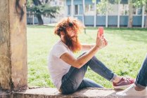 Молодий чоловічий хіпстер з рудим волоссям і бородою фотографує друга на смартфоні в парку — стокове фото