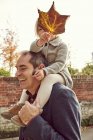 Daughter holding autumn leaf while sitting on father shoulders — Stock Photo