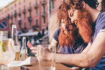 Young male hipster twins with red hair and beards reading smartphone texts at sidewalk bar — Stock Photo