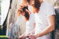 Gemelos hipster masculinos jóvenes con barbas rojas apoyadas en mensajes de texto de pared en teléfonos inteligentes - foto de stock