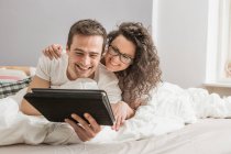Пара лежить на ліжку і використовує цифровий планшет — стокове фото