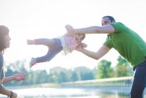 Mid adult couple swinging toddler daughter at riverside — Stock Photo