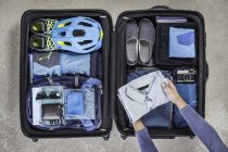 Vista aérea de las manos del hombre maleta de embalaje con botas de paseo, casco de bicicleta, mochila, cámara retro y camisa azul - foto de stock