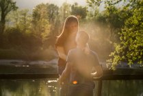 Пара біля річки, молода жінка сидить на паркані, стикається з молодим чоловіком, посміхаючись — стокове фото