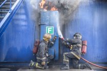 Bomberos atacando llamas detrás de la puerta de acero en instalaciones de entrenamiento de simulación de incendios - foto de stock