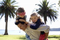 Mature man giving piggy back to daughters in coastal park, New Zealand — Stock Photo