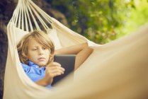 Junge liegt in Gartenhängematte und surft mit digitalem Tablet — Stockfoto