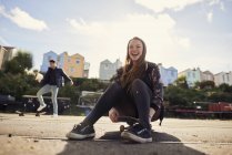 Две подруги, болтающиеся на улице, молодая женщина, сидящая на скейтборде, смеется, Бристоль, Великобритания — стоковое фото