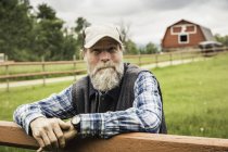 Бородатий чоловік на фермі спирається на паркан, дивлячись на камеру посміхаючись — стокове фото