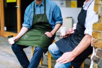 Senior craftsman drinking coffee and chatting to young man outside workshop, mid section — Stock Photo