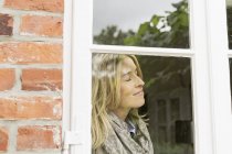 Mujer adulta por ventana con los ojos cerrados, sonriendo - foto de stock