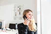 Young woman in office using smartphone to make telephone call looking away smiling — Stock Photo