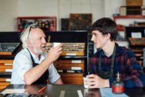 Senior-Handwerker trinkt Kaffee und plaudert mit jungem Handwerker in Buchkunstwerkstatt — Stockfoto