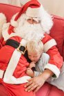 Man sitting on sofa dressed as santa hugging boy — Stock Photo