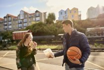 Joven compartiendo bolsa de papas fritas con un amigo, joven sosteniendo baloncesto, Bristol, Reino Unido - foto de stock