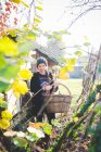 Blick durch Blätter einer jungen Frau im Garten mit Strickmütze und geflochtenem Korb, die in die Kamera lächelt — Stockfoto