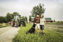 Пара з собакою на фермі перед трактором, дивлячись на камеру посміхаючись — стокове фото