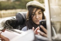Молодая женщина носит плоскую кепку в кабриолете с помощью смартфона — стоковое фото