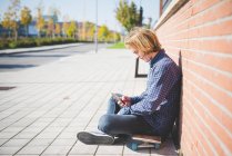 Молодой городской скейтбордист сидит на тротуаре и подбирает музыку для смартфонов — стоковое фото