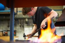 Young male trainee blacksmith hammering red hot metal on workshop anvil — Stock Photo