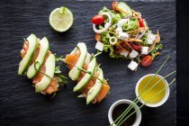 Smoked fish and avocado open sandwiches with salad and dipping sauces on slate, top  view — Stock Photo