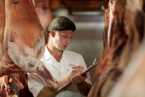 Carniceiro macho com prancheta inspecionando carne — Fotografia de Stock