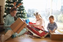 Хлопчик і сестри, сидячи на підлозі вітальні, відкривають різдвяні подарунки — стокове фото