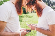 Молоді чоловічі хіпстерські близнюки з червоними бородами навпроти один одного смс на смартфонах — стокове фото