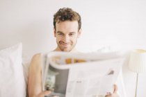Junger Mann liegt grinsend im Bett, während er Zeitung liest — Stockfoto