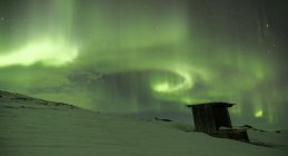 Nordlichter am Himmel über schneebedecktem Hügel und Hütte — Stockfoto