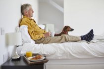 Senior man enjoying breakfast in bed with dog — Stock Photo