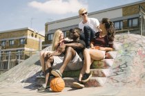 Cuatro amigos de baloncesto masculino y femenino sentados charlando en el parque de skate de la ciudad - foto de stock