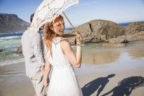Пара держащихся за руки, идущих по береговой линии, держащих зонтик, оглядывающихся через плечо на улыбающуюся камеру — стоковое фото