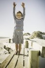 Portrait of young boy standing on wooden walkway at beach, arms raised — Stock Photo