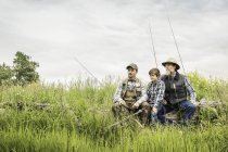 Multi generation family holding fishing rods sitting on log in field looking away — Stock Photo