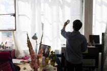 Rückwärtige Silhouette eines Mannes, der Druck auf das sonnenbeschienene Fenster im Druckereistudio hochhält — Stockfoto