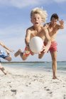 Мальчик прыгает в воздухе с мячом для регби, преследуемый братом и отцом на пляже, Майорка, Испания — стоковое фото
