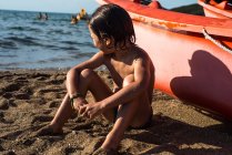 Boy sitting on beach by boat looking away — Stock Photo