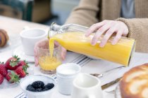 Vista cortada de mãos femininas derramando suco de laranja de garrafa em vidro — Fotografia de Stock
