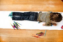 Visão aérea do menino deitado em cima e desenhando em papel longo — Fotografia de Stock
