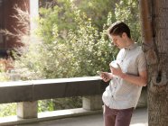 Junger Mann im Freien, an Baum gelehnt, Smartphone in der Hand, Imbissbecher — Stockfoto