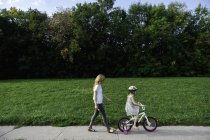 Jovencita montando su bicicleta seguida de madre - foto de stock
