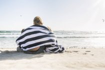 Vista posteriore di padre e figli seduti sulla spiaggia avvolti in una coperta — Foto stock