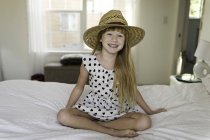 Menina sentada na cama sorrindo, usando chapéu de palha — Fotografia de Stock
