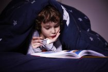 Junge unter Bettdecke liest Buch bei Fackelschein — Stockfoto