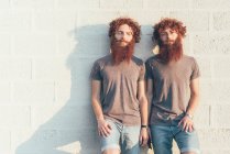 Retrato de gemelos adultos idénticos con pelo rojo y barba contra la pared - foto de stock